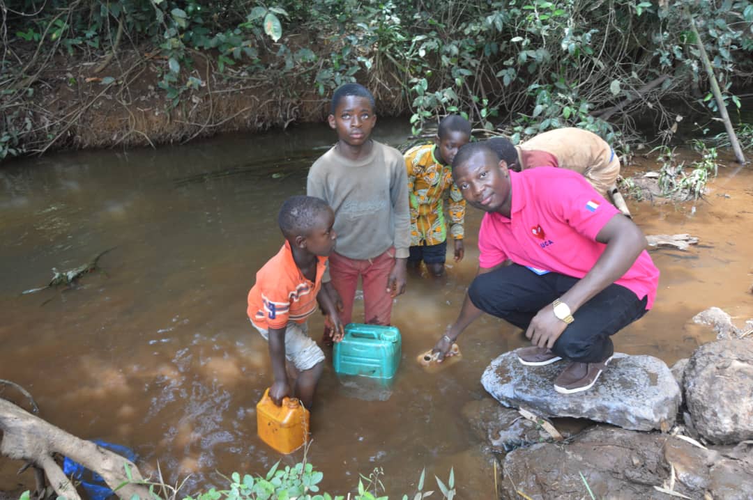 The Director of Un Cœur Qui Agit stands with children collecting muddy water from a stream in a rural community in Cameroon, highlighting the urgent need for access to safe drinking water.